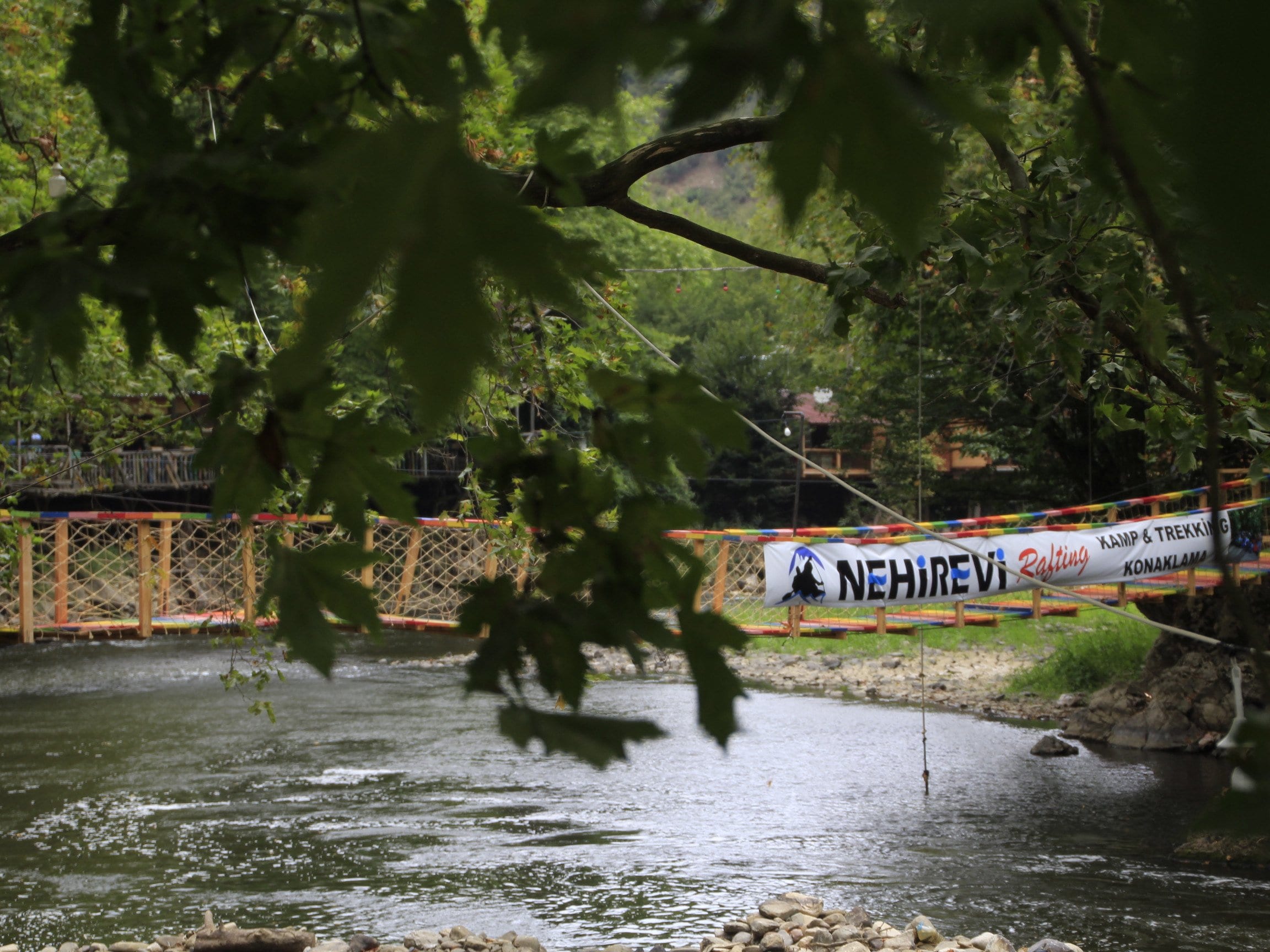 Çınar yapraklarının arasından görünen Melen Çayı asma köprüsü ve köprüye asılı Nehir Evi Rafting pankartı