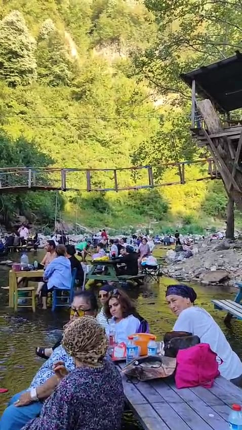 Nehir kenarı kamp alanında asma köprüden görülen piknik masaları ve suda serinleyen ziyaretçiler