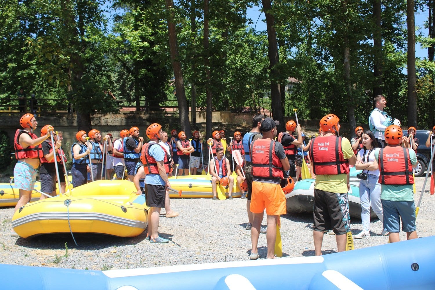 Melen Çayı rafting turu öncesi çakıl kıyıda sarı ve mavi botların yanında rafting ekipmanı ve kürek dağıtılan katılımcı grubu