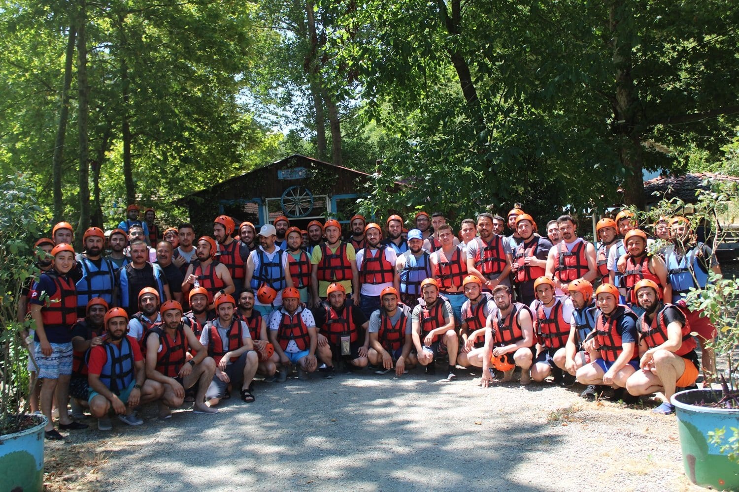 Kurumsal rafting organizasyonunda ahşap kulübe önünde can yeleği ve kaskla toplu poz veren kalabalık şirket ekibi