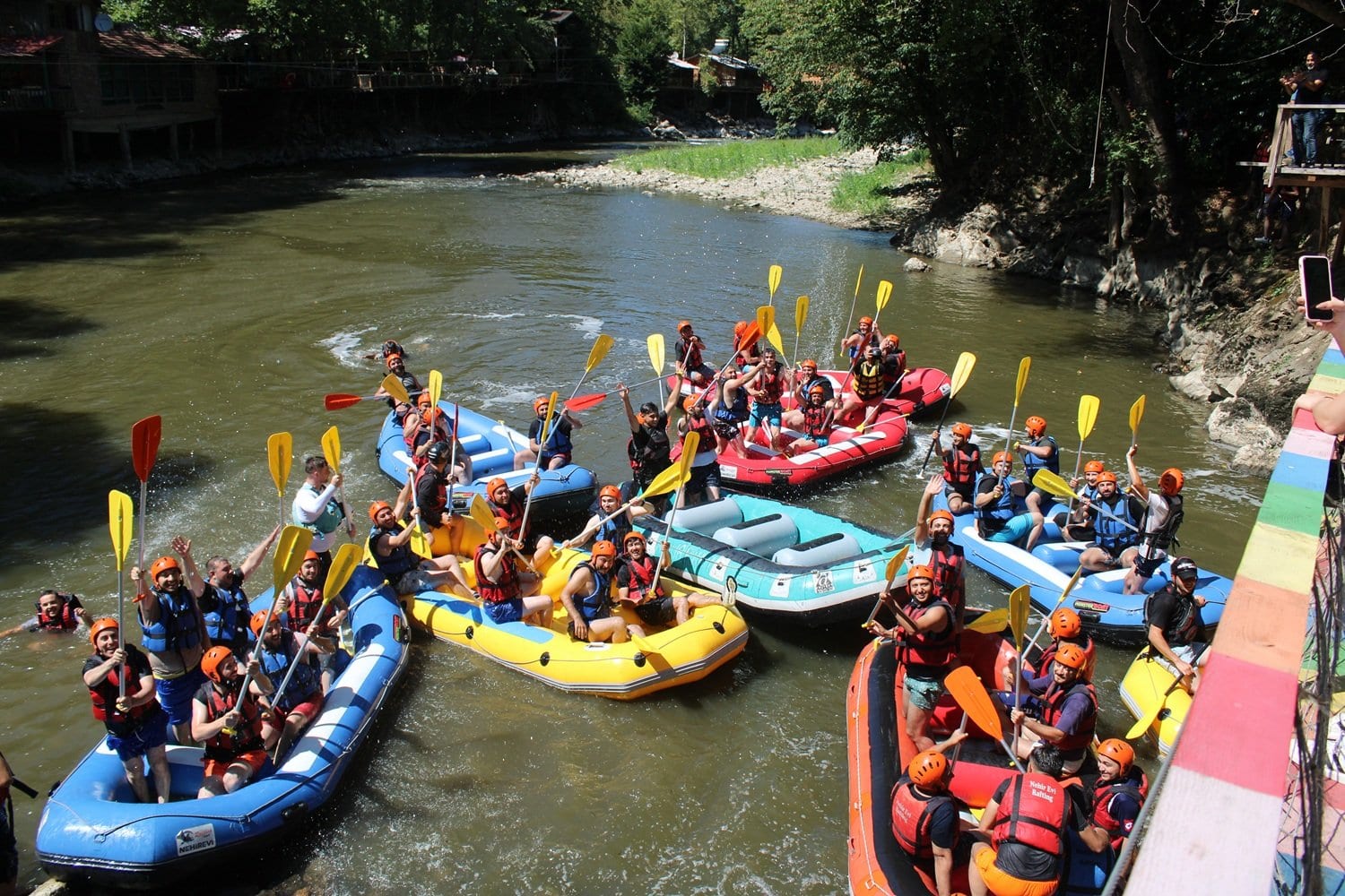 Renkli asma köprüden bakıldığında Melen Çayı üzerinde toplanan rafting bot filosu ve kürekleri kaldıran kurumsal grup