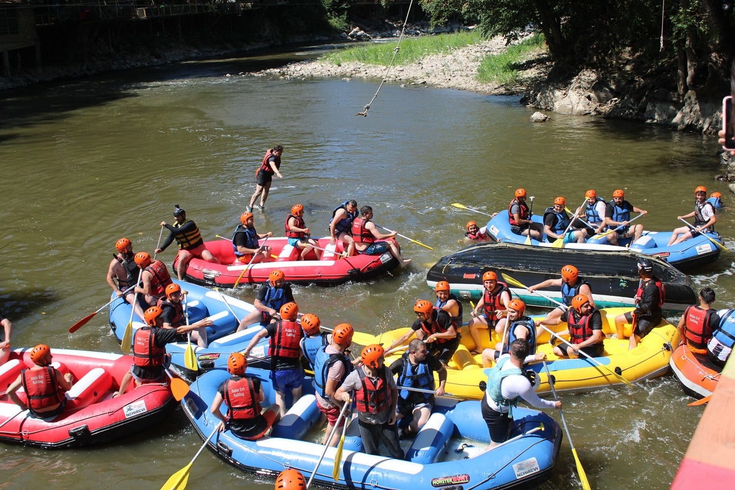 Kurumsal rafting organizasyonunda nehrin ortasında yan yana toplanan onlarca rafting botu ve üstlerinden geçen ip hattı