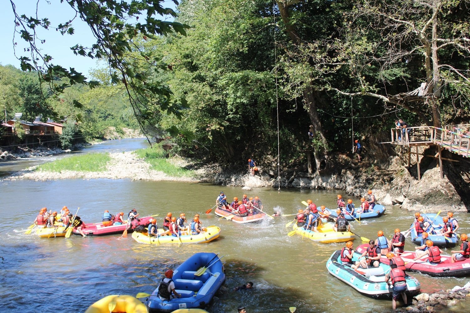 Nehir kenarındaki ahşap terasların önünden görünen Melen Çayı ve kurumsal rafting grubunun dağılmış bot filosu