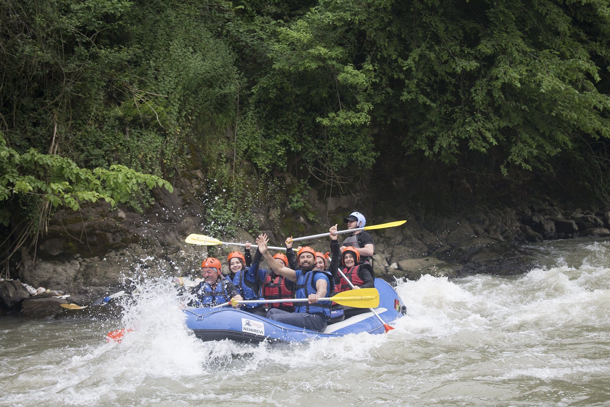 Melen Çayı rafting parkurunda kürekleri havaya kaldırıp sevinen mavi bottaki katılımcılar ve yandan gelen köpük dalgası