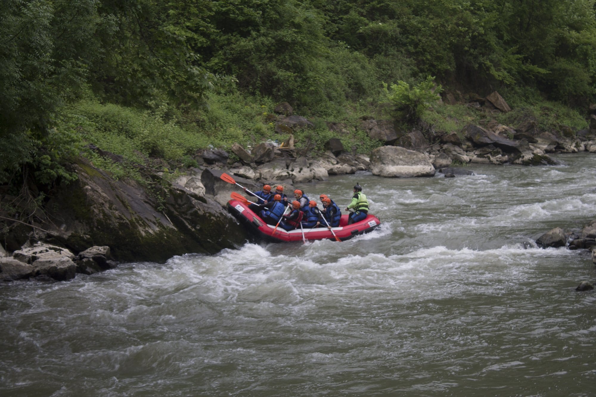 Düzce rafting turunda kayalık yamacın dibinden kırmızı botla köpüklü akıntıya giren ekip ve dümendeki rehber