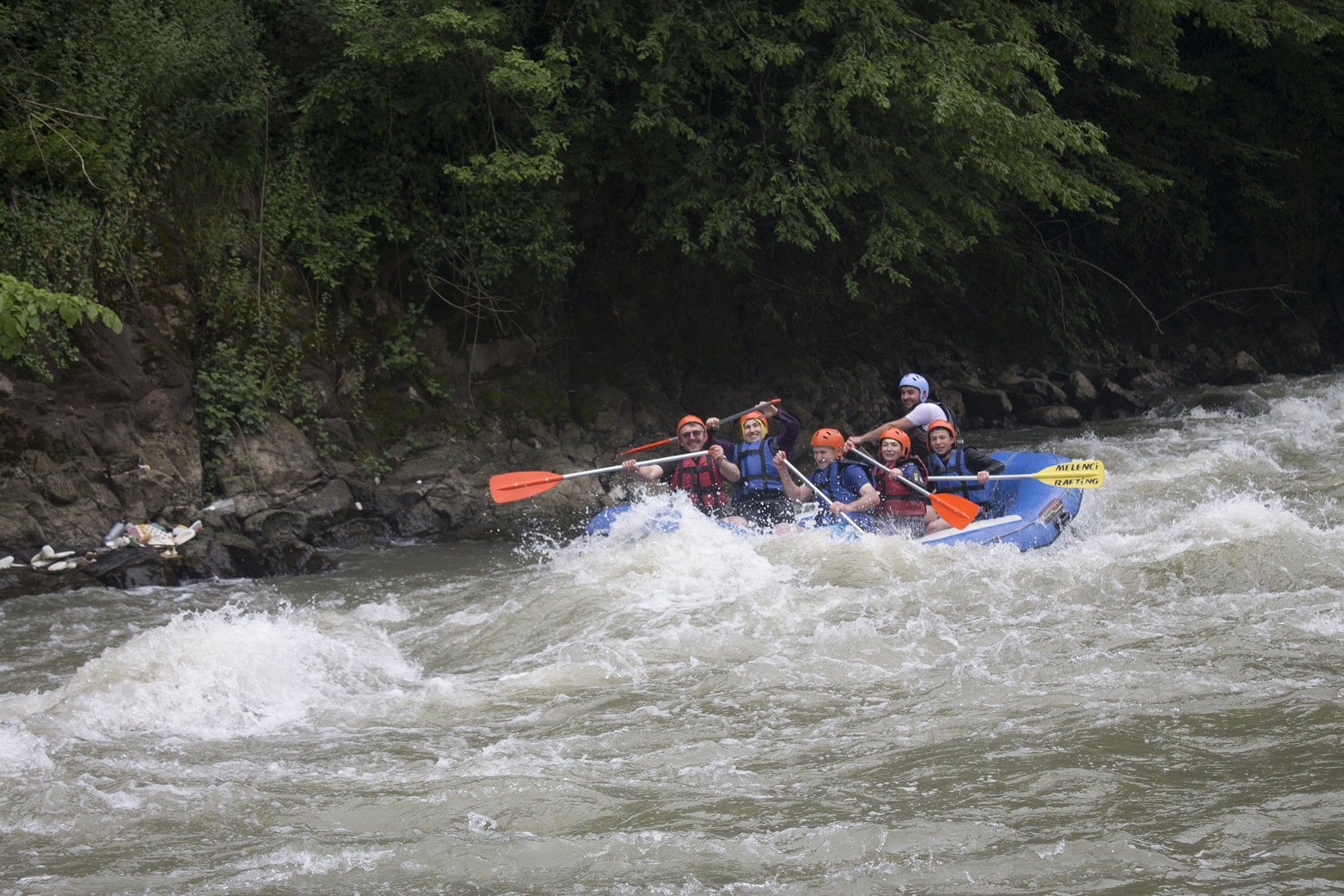 Melen Çayı rafting parkurunda duran dalganın yanından süzülen mavi bot ve kürek çeken turuncu kasklı katılımcılar