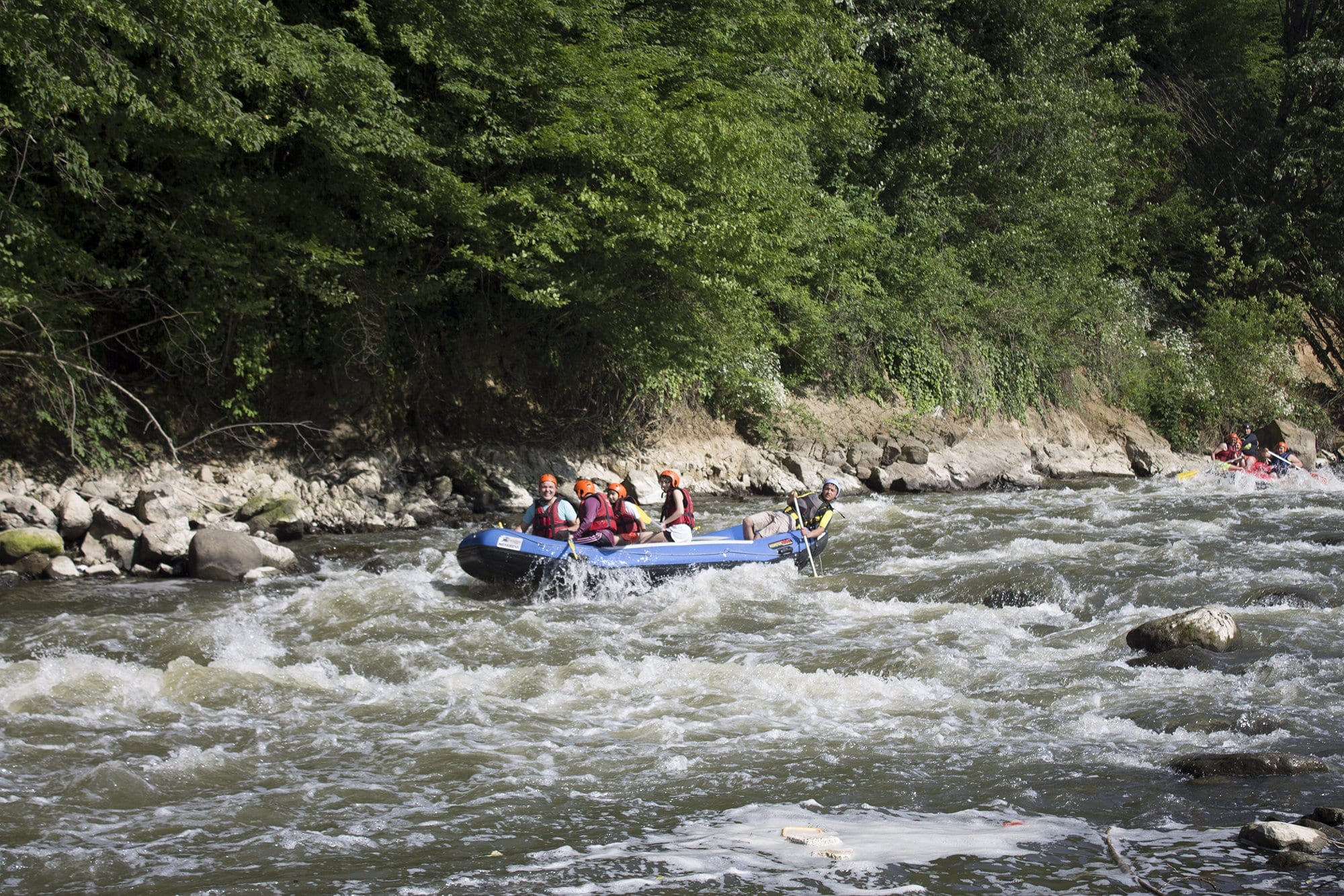 Cumayeri rafting parkurunda iri taşlarla kaplı kıyı boyunca ilerleyen mavi bot ve arkadan gelen ikinci rafting botu